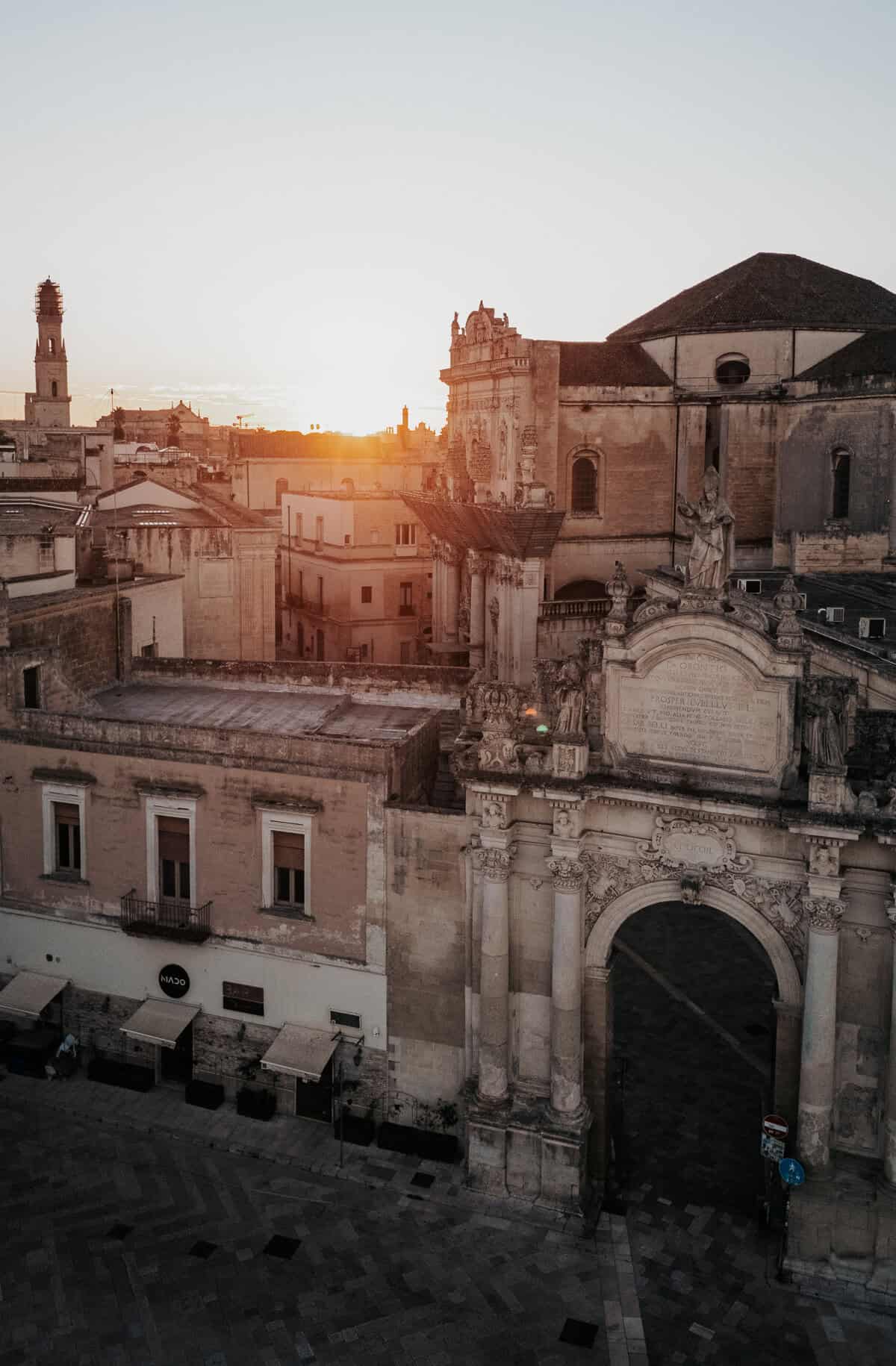 A stunning sunrise over the historic city of Lecce, Italy, casting a golden glow on the Baroque architecture. The ornate Porta Napoli arch stands prominently in the foreground, adorned with statues and intricate carvings, while the dome of a historic church rises behind it. The warm tones of the limestone buildings contrast beautifully with the soft morning sky.