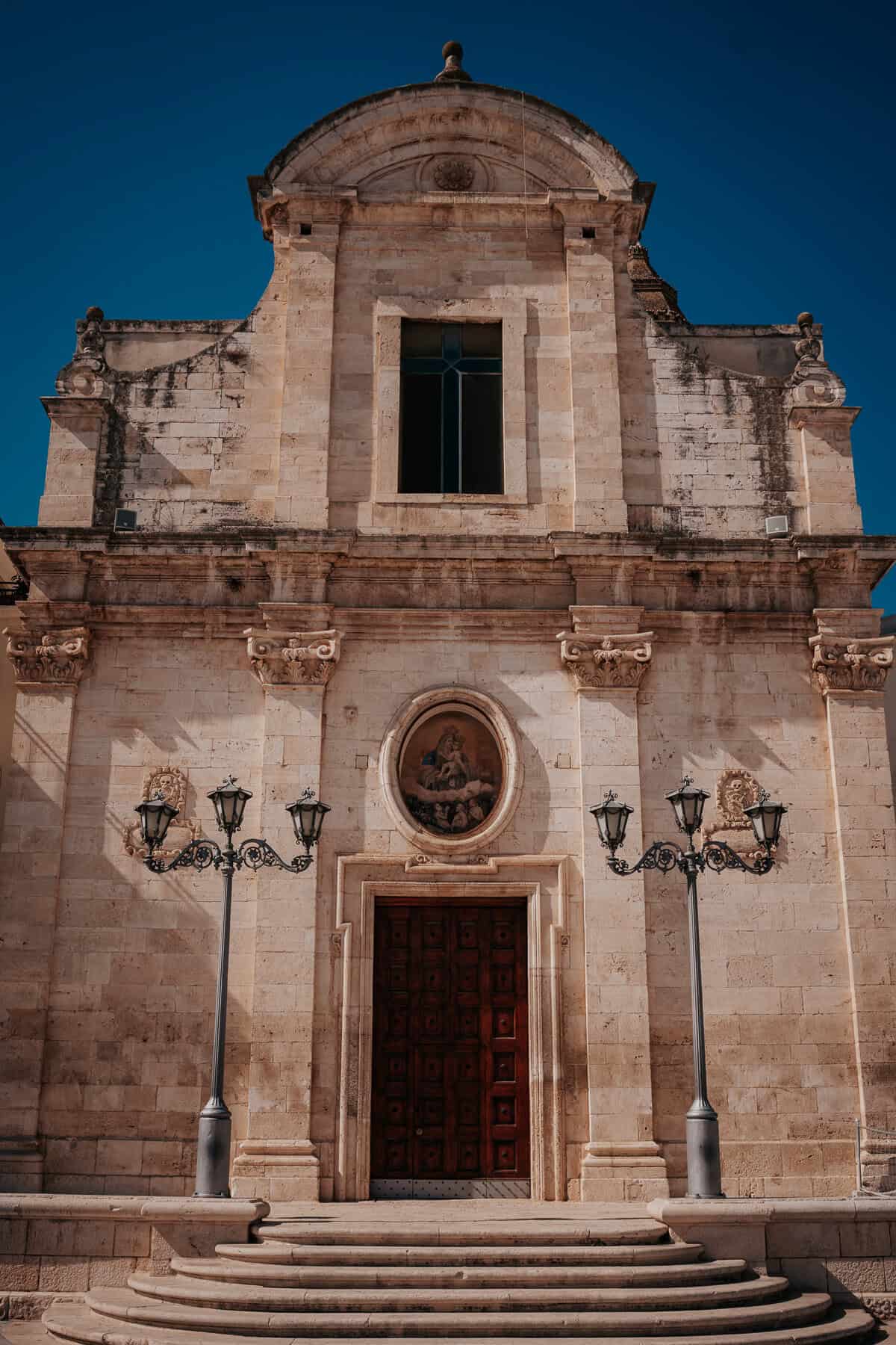 A historic stone church in Puglia with an ornate arched doorway, a large wooden door, and classical architectural details. Two vintage-style lampposts frame the steps leading up to the entrance beneath a deep blue sky.