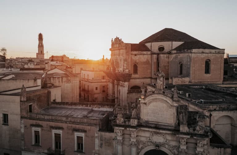 Sunset over the historic rooftops and stone buildings of Lecce, Italy, with warm golden light hitting a Baroque church and bell tower. The architecture glows in soft evening light, capturing the charm of southern Italy.
