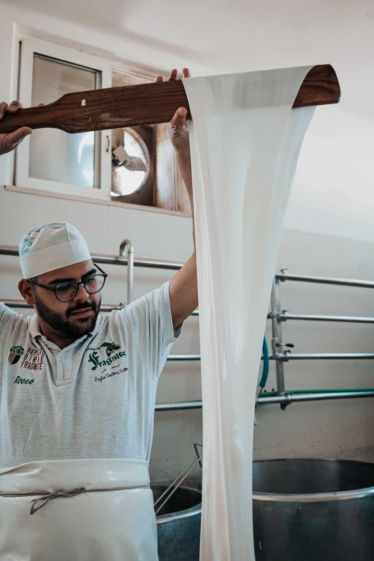 A man named Rocco stretches a long ribbon of mozzarella curd with a wooden paddle, demonstrating the traditional cheese-making process in a dairy room.