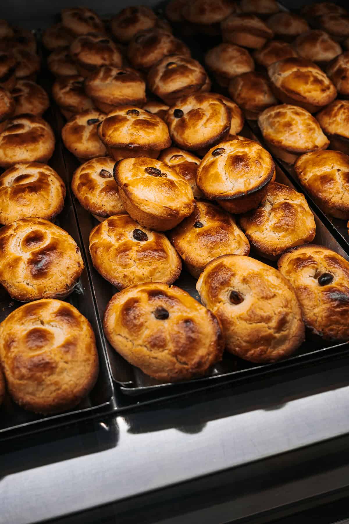 Rows of golden-brown pasticciotti pastries, each topped with a single raisin, fill trays at a bakery counter, fresh from the oven and ready for sale.