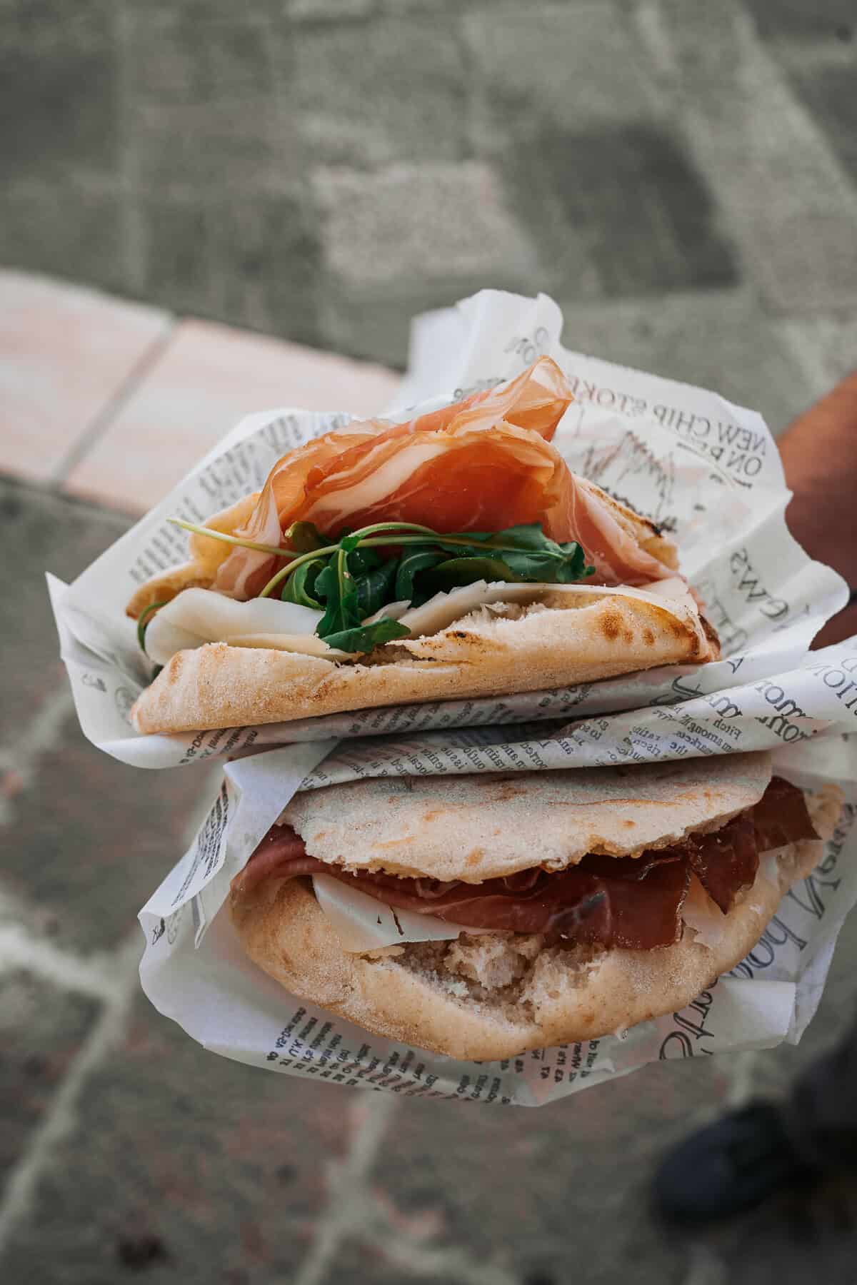 Two freshly made Italian sandwiches wrapped in paper, stuffed with cured meats, arugula, and cheese, held up against a stone street background.