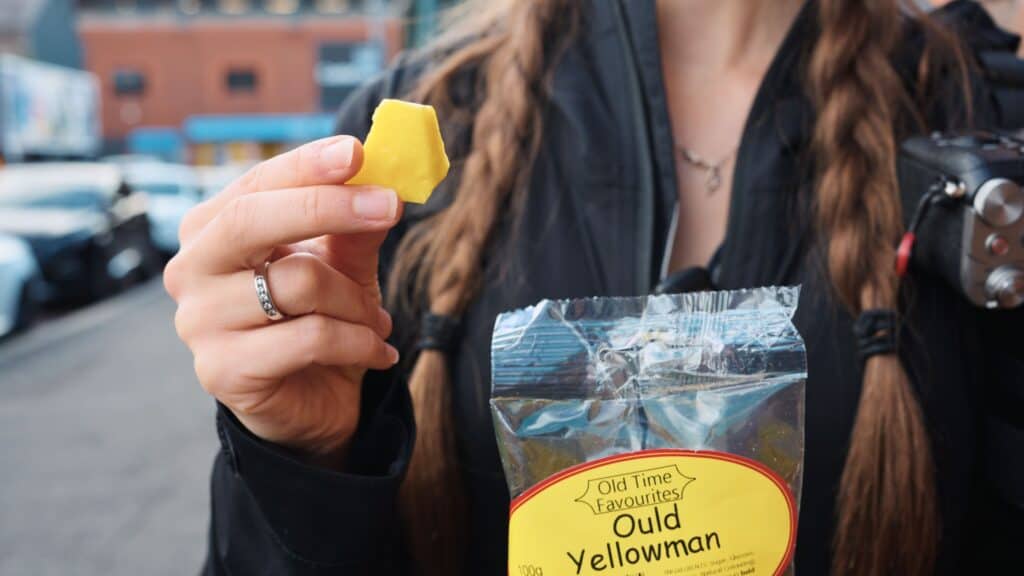 Hand holding a bright yellow cube of candy from a bag labeled “Old Time Favourites Ould Yellowman.” The honeycomb toffee sweet is a traditional Scottish candy often found at markets and sweet shops.