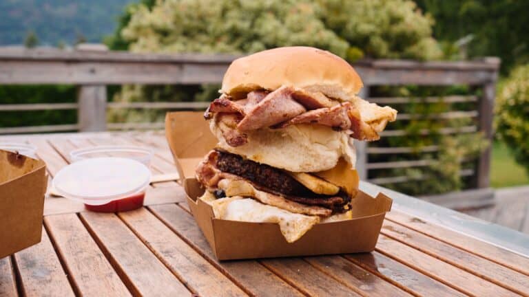Tall breakfast roll stacked with fried egg, sausage patty, bacon, and sliced meat inside a soft white bun served in a takeaway tray on an outdoor table. The towering sandwich highlights hearty traditional Scottish food often enjoyed as a filling breakfast.