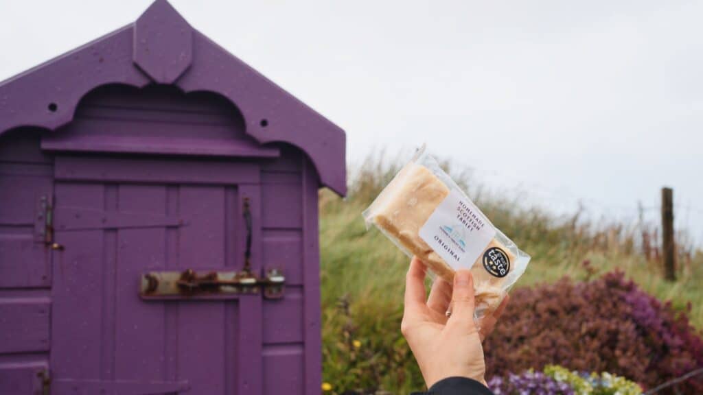 Hand holding a packaged tablet candy labeled “Homemade Scottish Tablet Original” in front of a purple wooden seaside hut and grassy coastal landscape. The crumbly fudge like sweet is a classic traditional Scottish food treat.