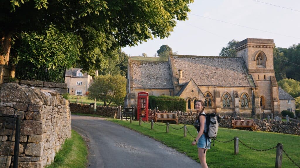 A traveler with a backpack walks along a quiet village road past a historic stone church and a red telephone box. This weekend in the Cotswolds scene captures the charm of a traditional English village.