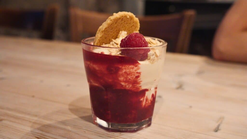Glass dessert cup layered with cream, oats, raspberry sauce, and a fresh raspberry topped with a biscuit. The layered dessert resembles cranachan, a sweet traditional Scottish food made with berries, cream, and oats.