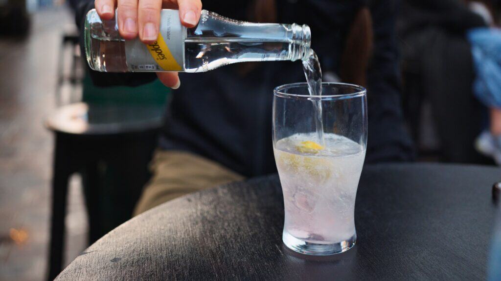 Person pouring clear soda water from a small bottle into a glass filled with ice and a lemon slice at an outdoor table. Sparkling water is often used as a mixer with Scotch whisky, a key part of Scottish food and drink culture.