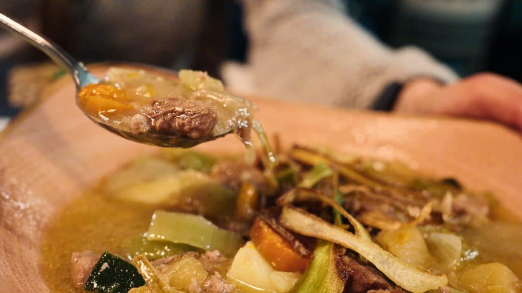 Spoon lifting a bite of hearty beef stew with vegetables including potatoes, carrots, and leeks from a bowl. The slow cooked dish reflects comforting traditional Scottish food often served in pubs.