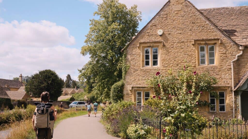 A traveler walks past honey colored stone cottages and blooming flowers along a peaceful village street. This weekend in the Cotswolds image shows the picturesque countryside setting.