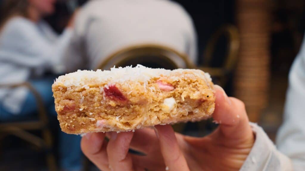 Hand holding a thick dessert bar topped with coconut flakes and filled with pink and white pieces. The sweet traybake resembles a bakery style treat commonly found in Irish cafés.