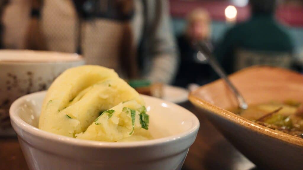 Bowl of creamy mashed potatoes mixed with green herbs sitting beside a dish of stew on a wooden table. The buttery mash resembles traditional Scottish tatties served with hearty meals.