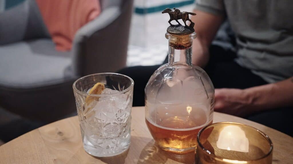 A glass of cocktail sits beside a decorative bottle with a horse stopper on a small table in a dimly lit bar. This best restaurants in the Cotswolds scene captures a cozy drink setting.