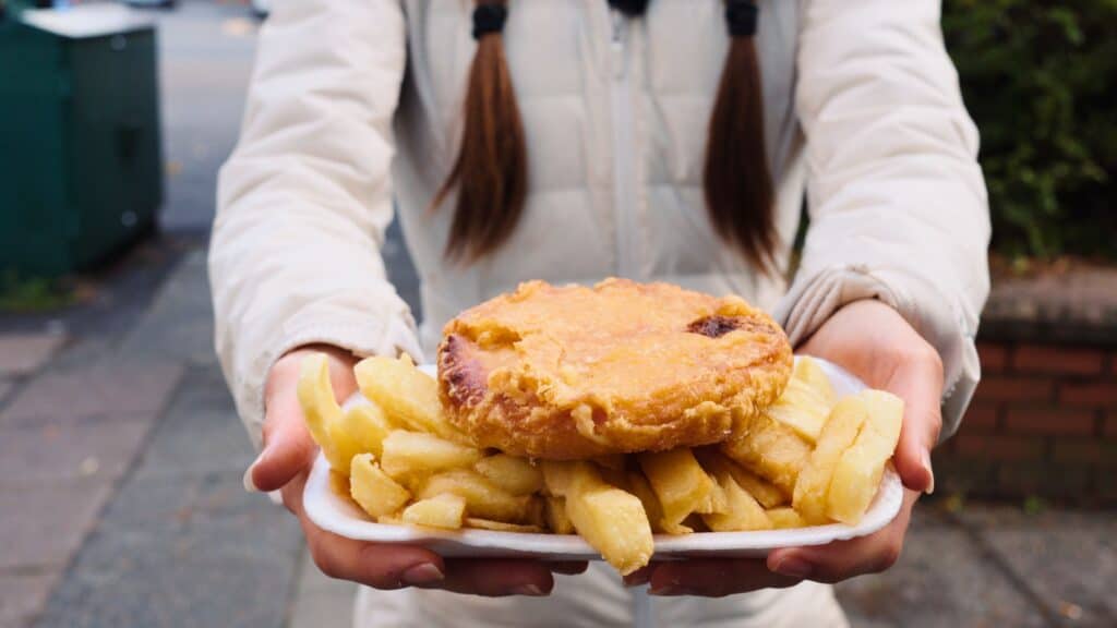 Person holding a tray of thick cut chips topped with a golden meat pie outdoors. The takeaway pie and chips are a classic comfort meal in Scottish food culture.